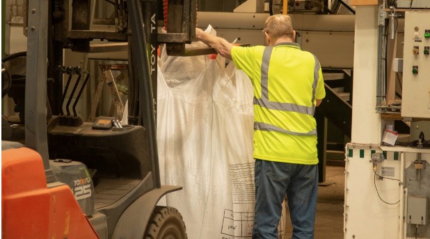 A man in a yellow high-visibility vest attaches a bag of seed to a fork lift